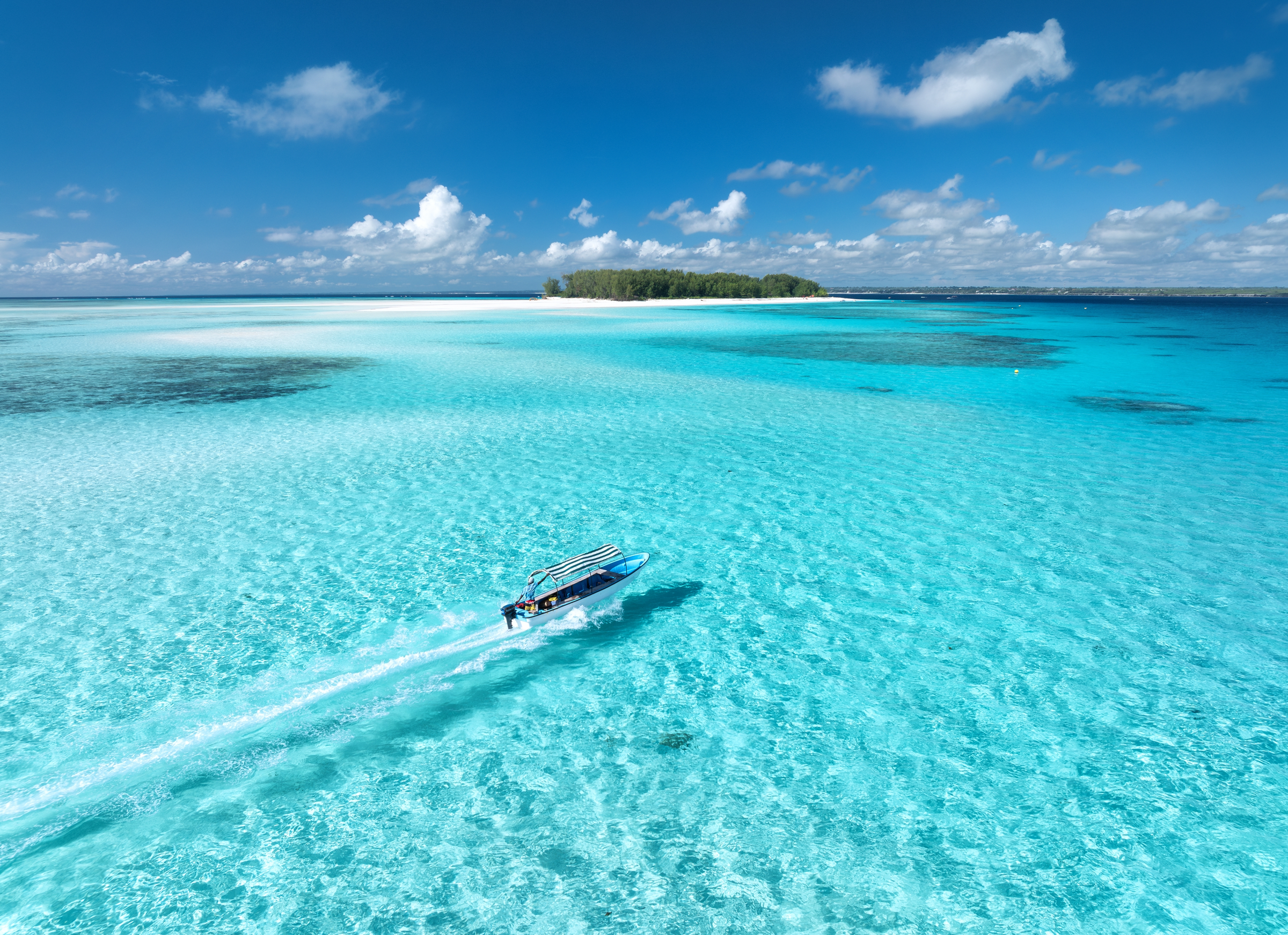 Aerial view of floating boat in transparent azure water on sunny summer day. Mnemba island, Zanzibar. Top view of yacht, sandbank in low tide, clear blue sea, sand, sky with clouds. Motorboat in ocean