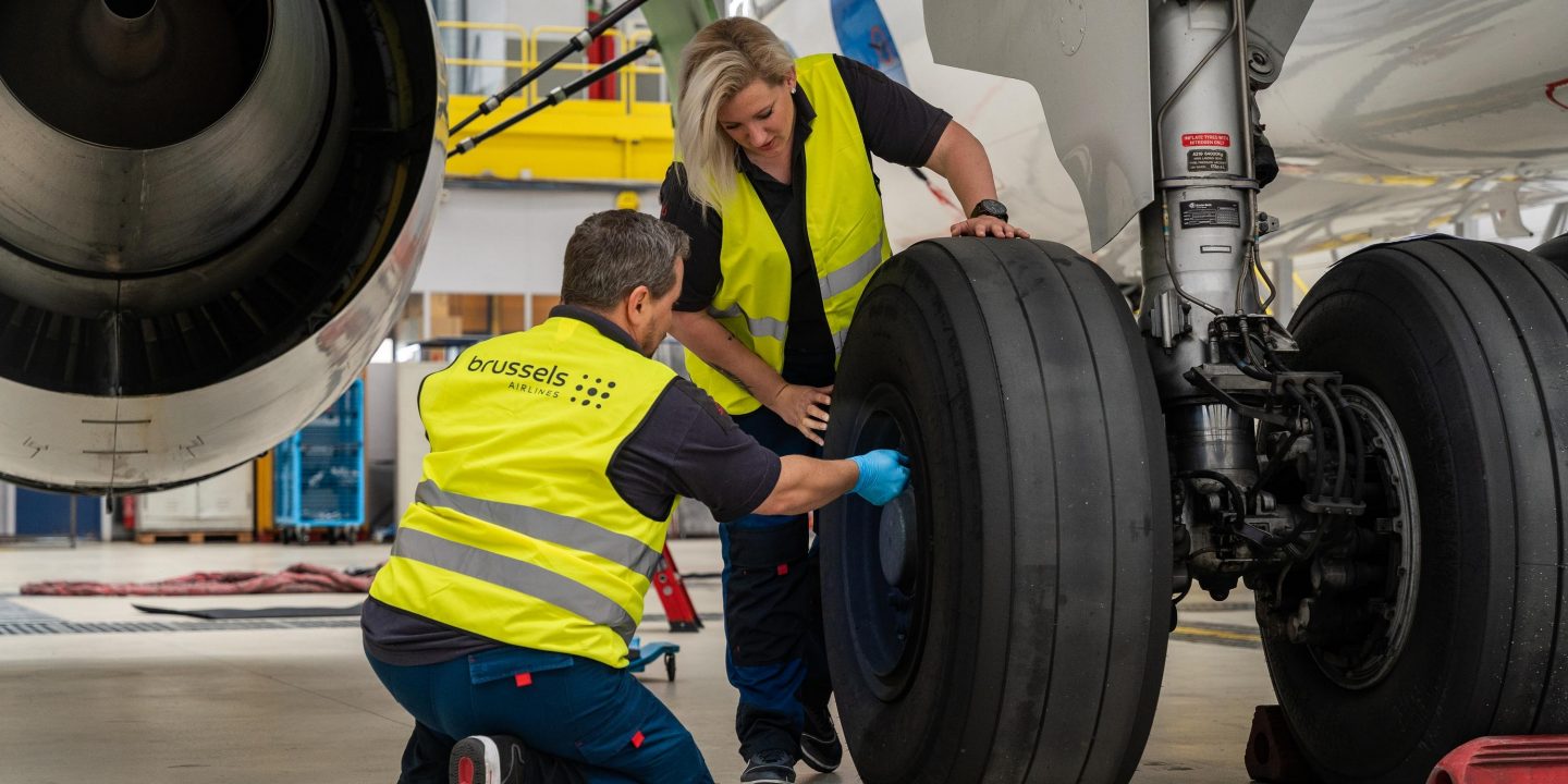 Two Brussels Airlines technicians in high-visibility vests inspect the landing gear of an aircraft in a maintenance hangar.