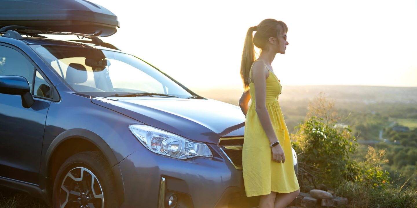 Yong happy woman standing near her car enjoying sunset view of summer nature. Travel and getaway concept.