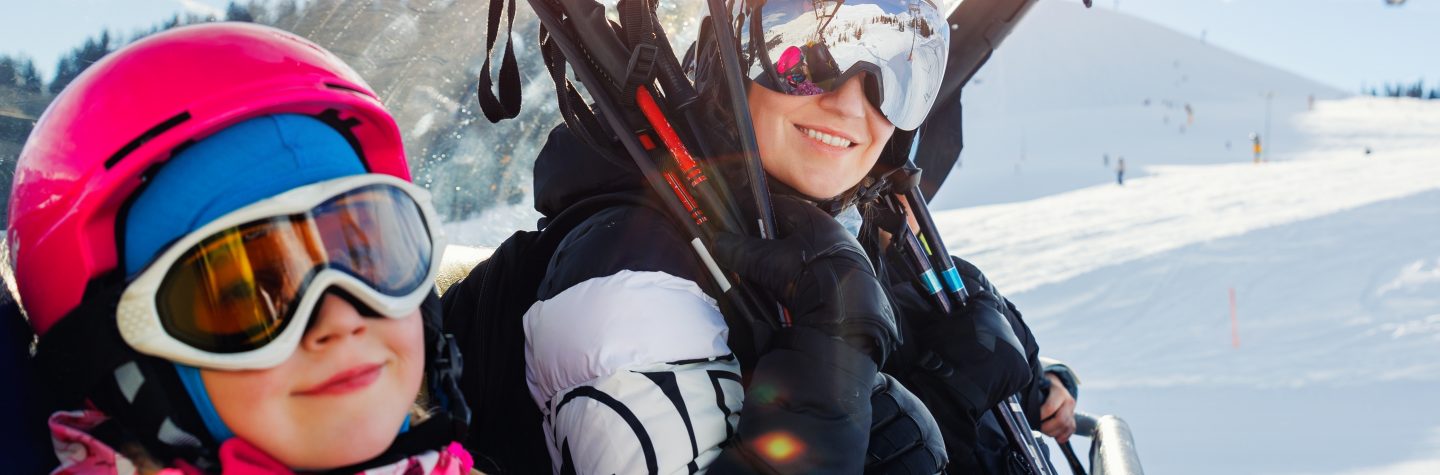 Cute little girl with mom in helmet and goggles having fun sitting on ski lift chair enjoying winter ski vacations in alpine austrian mountains. Happy family portrait winter sport holidays