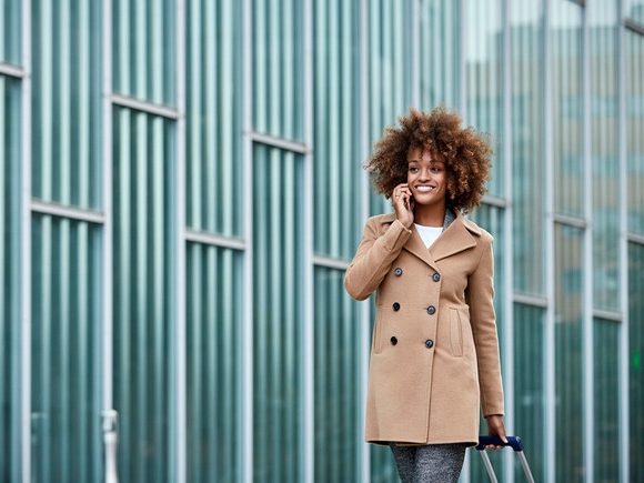 Young businesswoman with luggage talking on mobile phone. Confident female professional is walking by building. She is wearing overcoat during business trip.