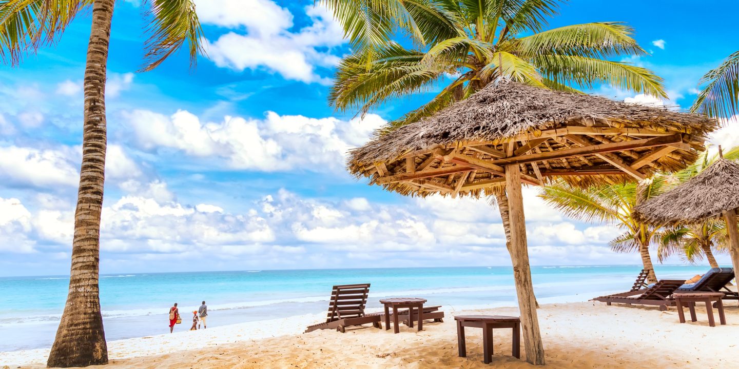 Sun loungers under umbrella and palms on the sandy beach by the ocean and cloudy sky. Vacation background. Idyllic beach landscape in Diani beach, Kenya, Africa.